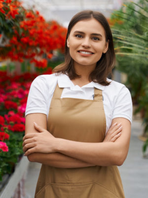 Beautiful young woman with brown hair holding hands crossed while posing at greenhouse. Smiling florist in beige apron working with various colorful flowers.
